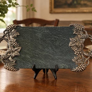 Puede incluir: Una bandeja rectangular de pizarra verde oscuro con asas metálicas ornamentadas en ambos lados. Las asas están esculpidas con racimos de uvas y hojas de parra detalladas. La bandeja se sostiene sobre un pequeño soporte de plástico negro.