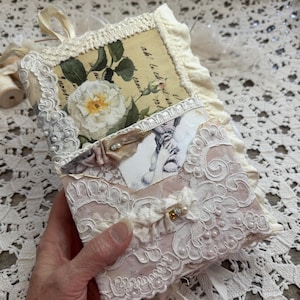 May include: A handmade journal featuring a cream-colored fabric cover adorned with white lace, delicate pearl beads, and a ruffled edge. The front displays a vintage-style floral print with a white rose and a small pocket containing a black and white image.