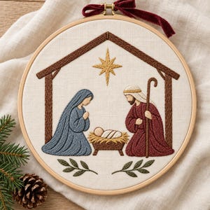 May include: A circular wooden embroidery hoop containing a fabric panel with a stitched nativity scene. The design features a brown stable outline, a yellow star, and figures in blue and red robes surrounding a baby in a manger.