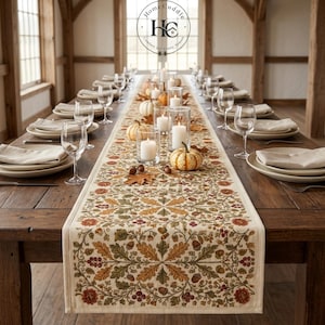 May include: A long rectangular table runner featuring a detailed autumnal pattern of oak leaves, acorns, and berries in shades of orange, brown, and green on a cream background, centered on a wooden dining table with place settings and candles.