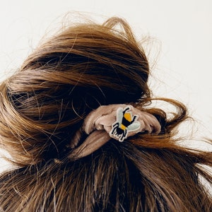 Pu&ograve; includere: Un elastico per capelli marrone con una piccola toppa ricamata a forma di ape. L'ape ha un corpo giallo, strisce nere e ali azzurre. L'elastico &egrave; realizzato in un tessuto morbido e strutturato e viene indossato in uno chignon.