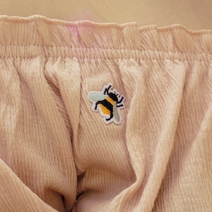 Peut inclure: Un haut rose clair à épaules dénudées avec un petit écusson brodé d'abeille. Le haut a un col à volants et une texture en velours côtelé. Un collier en or est également visible.