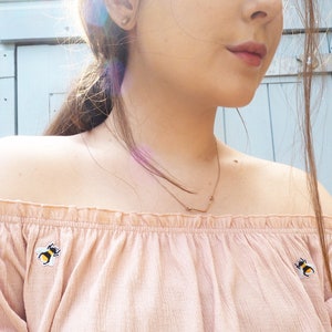 Pu&ograve; includere: Un top rosa chiaro con spalle scoperte con toppe a forma di ape ricamate. Si indossa una collana color oro con un ciondolo a forma di V. Il top &egrave; realizzato in un tessuto morbido e strutturato.