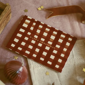 May include: A Christmas card with a red and white checkered pattern and the words "HAPPY CHRISTMAS" in gold. The card is on a pink surface with gold star-shaped confetti, a brown ribbon, and a red ornament.