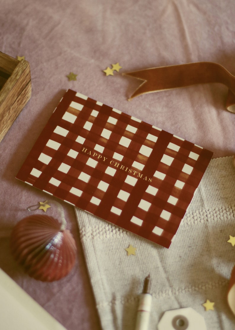 May include: A Christmas card with a red and white checkered pattern and the words "HAPPY CHRISTMAS" in gold. The card is on a pink surface with gold star-shaped confetti, a brown ribbon, and a red ornament.
