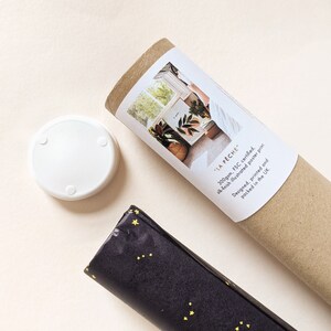 May include: A white coaster with three small feet, a rolled-up poster, and a wrapped package are shown from above. The poster is in a brown tube with a label that reads "LA P&Ecirc;CHE". The package is wrapped in black paper with yellow star accents.