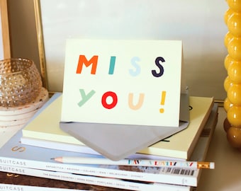 May include: A greeting card with the words "MISS YOU!" in colorful block letters. The card is on top of a stack of books and magazines, with a gray envelope. A pencil rests on the magazines.
