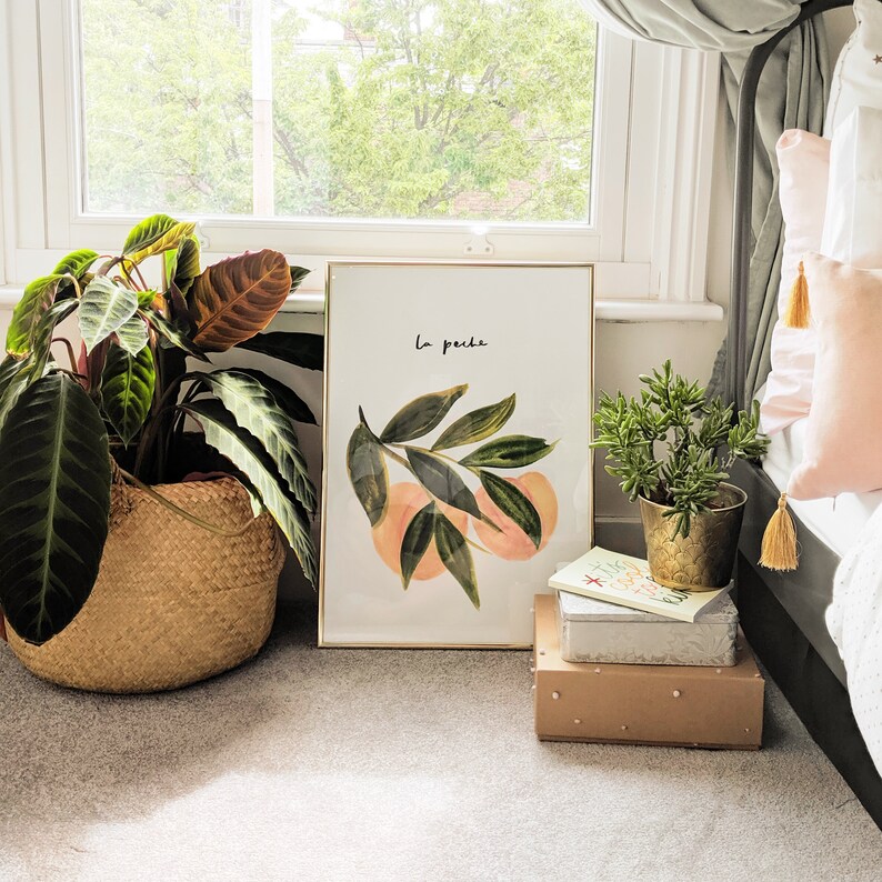 May include: A framed art print with a gold border, displaying a watercolour illustration of peaches and leaves, with the text "La peche". A potted plant in a woven basket and a small potted plant are also visible.