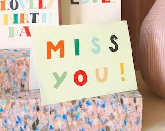 May include: A greeting card with the words "MISS YOU!" in colorful block letters. The card is light green and sits on a colorful foam block. Other cards with text are visible in the background, along with a pink vase.