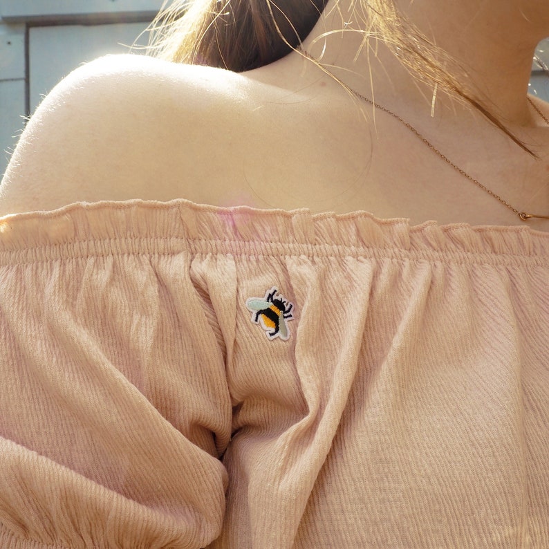 Pu&ograve; includere: Un top rosa chiaro con spalle scoperte con una piccola toppa ricamata a forma di ape. Il top ha uno scollo a balze e una trama a coste. &Egrave; visibile anche una collana d'oro.