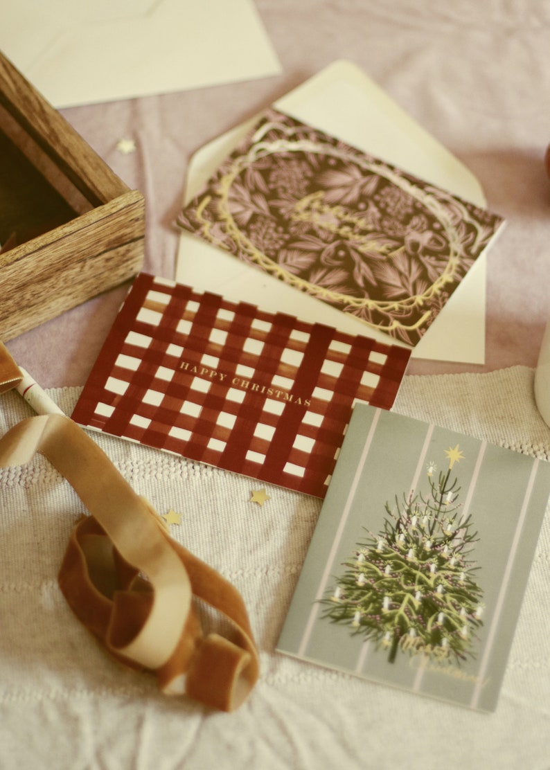 May include: Christmas cards with festive designs. One card features a red and white checkered pattern with "HAPPY CHRISTMAS" text. Another card has a Christmas tree illustration. A wooden box and a velvet ribbon are also present.