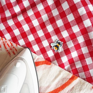 Pu&ograve; includere: Un ferro da stiro bianco &egrave; appoggiato su una tovaglia a scacchi rossa e bianca, accanto a una toppa a forma di ape colorata. La toppa raffigura un'ape nera e gialla con ali blu. La tovaglia &egrave; piegata su un tessuto fantasia.