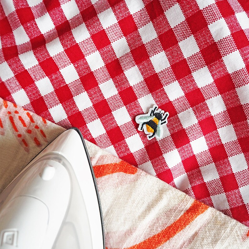 Pu&ograve; includere: Un ferro da stiro bianco &egrave; appoggiato su una tovaglia a scacchi rossa e bianca, accanto a una toppa a forma di ape colorata. La toppa raffigura un'ape nera e gialla con ali blu. La tovaglia &egrave; piegata su un tessuto fantasia.