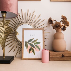 May include: A display of home decor items including a gold sunburst mirror, a framed watercolour print with the text "La p&ecirc;che", a pink candle, and a vase with dried flowers. A pink velvet lampshade is on the left.