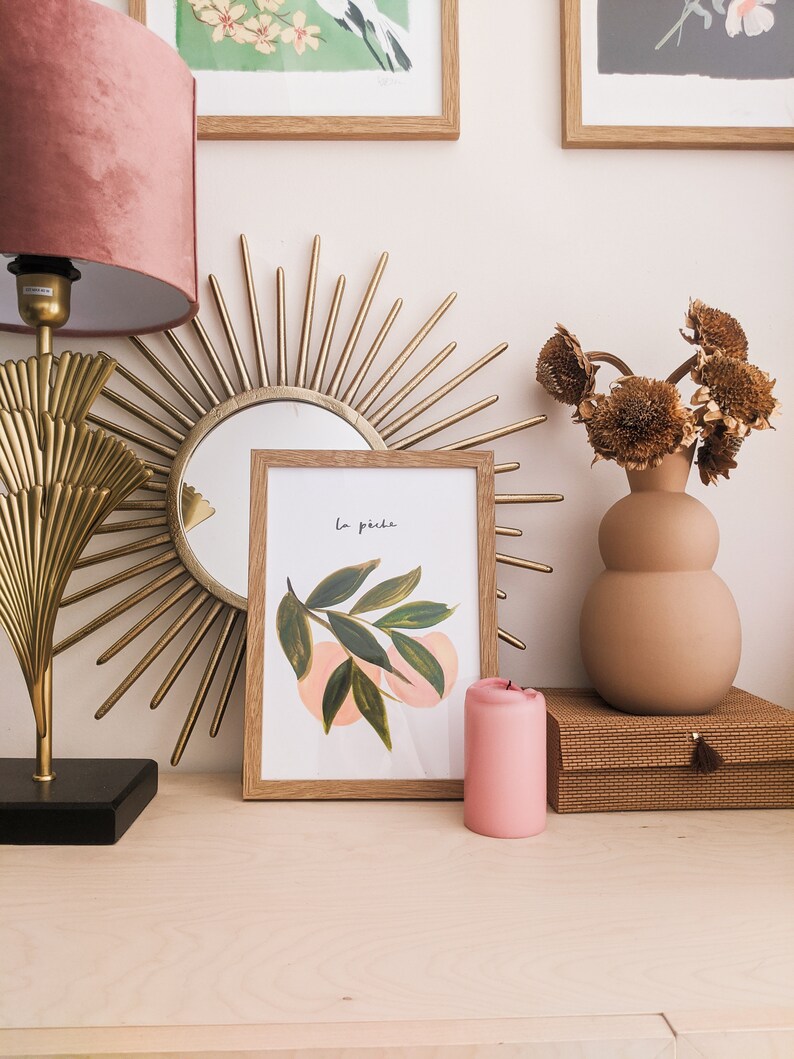 May include: A display of home decor items including a gold sunburst mirror, a framed watercolour print with the text "La p&ecirc;che", a pink candle, and a vase with dried flowers. A pink velvet lampshade is on the left.