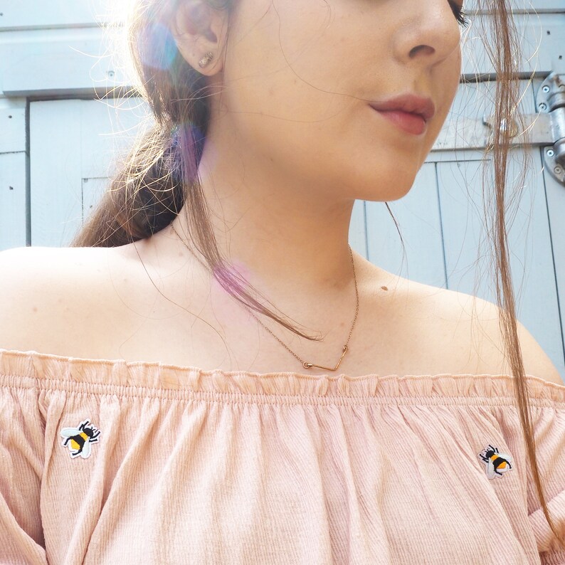 Pu&ograve; includere: Un top rosa chiaro con spalle scoperte con toppe a forma di ape ricamate. Si indossa una collana color oro con un ciondolo a forma di V. Il top &egrave; realizzato in un tessuto morbido e strutturato.