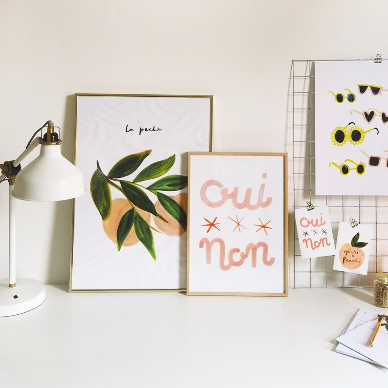 May include: A display of framed art prints and decorative items on a white surface. The prints showcase watercolour illustrations of a peach branch, sunglasses, and the words "Oui Non". A white desk lamp is also visible.