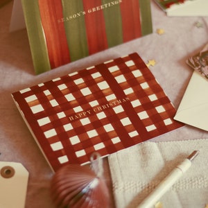 May include: Christmas greeting cards with various designs. One card features a red and white checkered pattern with "HAPPY CHRISTMAS" text. Other cards have striped and floral designs. A pen, envelopes, and decorative elements are also present.