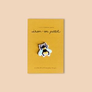 Pu&ograve; includere: Una toppa termoadesiva con un'ape ricamata con strisce nere e gialle, ali azzurre e dettagli bianchi. La toppa &egrave; su un cartoncino giallo senape con la scritta "iron-on patch" e "A Little Bit of Everyday Magic".