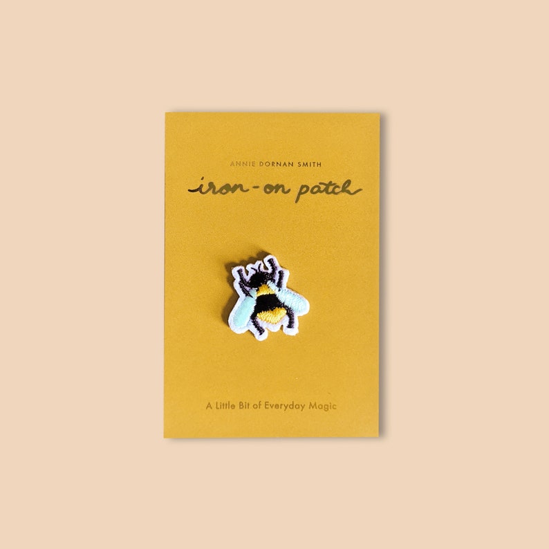 Pu&ograve; includere: Una toppa termoadesiva con un'ape ricamata con strisce nere e gialle, ali azzurre e dettagli bianchi. La toppa &egrave; su un cartoncino giallo senape con la scritta "iron-on patch" e "A Little Bit of Everyday Magic".