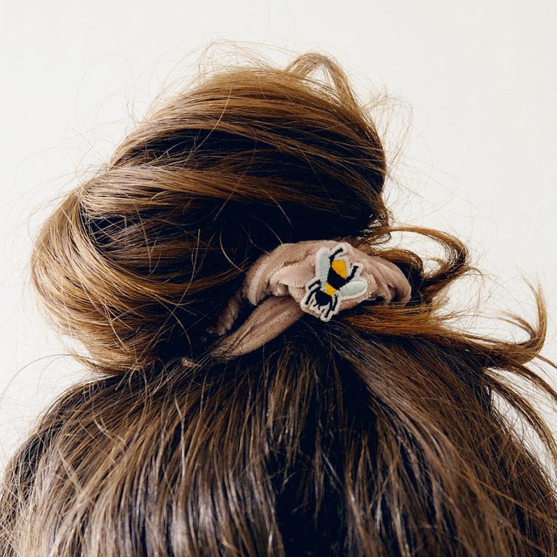 Pu&ograve; includere: Un elastico per capelli marrone con una piccola toppa ricamata a forma di ape. L'ape ha un corpo giallo, strisce nere e ali azzurre. L'elastico &egrave; realizzato in un tessuto morbido e strutturato e viene indossato in uno chignon.