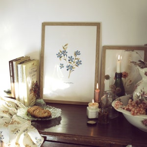 May include: A framed print of blue flowers with the word "meadows" in script. The print is on a dark wooden surface with books, candles, and a decorative plate with pastries. The scene evokes a vintage aesthetic.