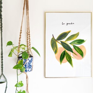 May include: A framed art print with a watercolour illustration of peaches and leaves, with the text "La peche". The artwork is in a gold frame. Two macram&eacute; plant hangers with potted plants are also visible.