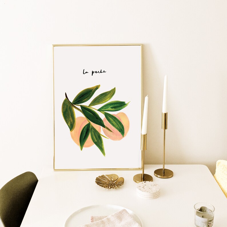 May include: A framed art print with a watercolour illustration of peaches and green leaves. The print has the text "La peche" in black script. Gold candle holders with white candles and a decorative dish are on a white table.