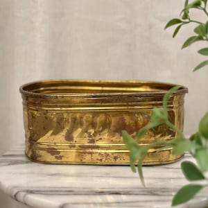 May include: An oval-shaped brass planter with a polished finish and visible patina spots. The container features a horizontal ribbed texture along the middle section and a smooth, rounded rim, providing a sturdy structure for holding items.