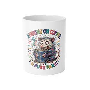 May include: A white ceramic mug featuring a colorful illustration of a wide-eyed opossum holding a blue coffee mug. The text reads RUNNING ON COFFEE & PURE PANIC, surrounded by lightning bolts, coffee beans, and stars in various bright colors.