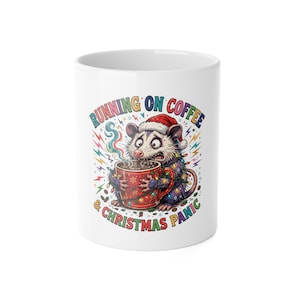 Peut inclure: Un mug en céramique blanc illustré d'un opossum aux yeux écarquillés portant un bonnet de Père Noël, entouré de guirlandes lumineuses et tenant une tasse rouge. Le texte indique RUNNING ON COFFEE & CHRISTMAS PANIC avec des éclairs.
