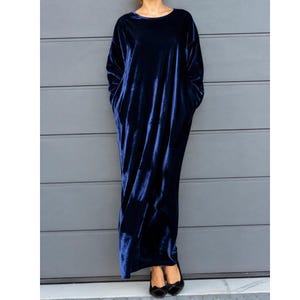 Könnte beinhalten: Ein bodenlanges Kleid mit langen Ärmeln aus dunkelblauem Samtstoff. Das Kleidungsstück verfügt über einen einfachen Rundhalsausschnitt, eine lockere, entspannte Silhouette und integrierte Seitentaschen, was für ein einheitliches Erscheinungsbild sorgt.