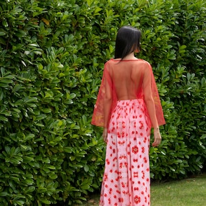 May include: A woman wearing a red sheer top over a pink and red cherry print maxi skirt. She is standing in front of a green hedge.