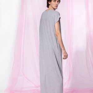 May include: A person wearing a long, gray, loose-fitting t-shirt dress standing in front of a pink sheer curtain.
