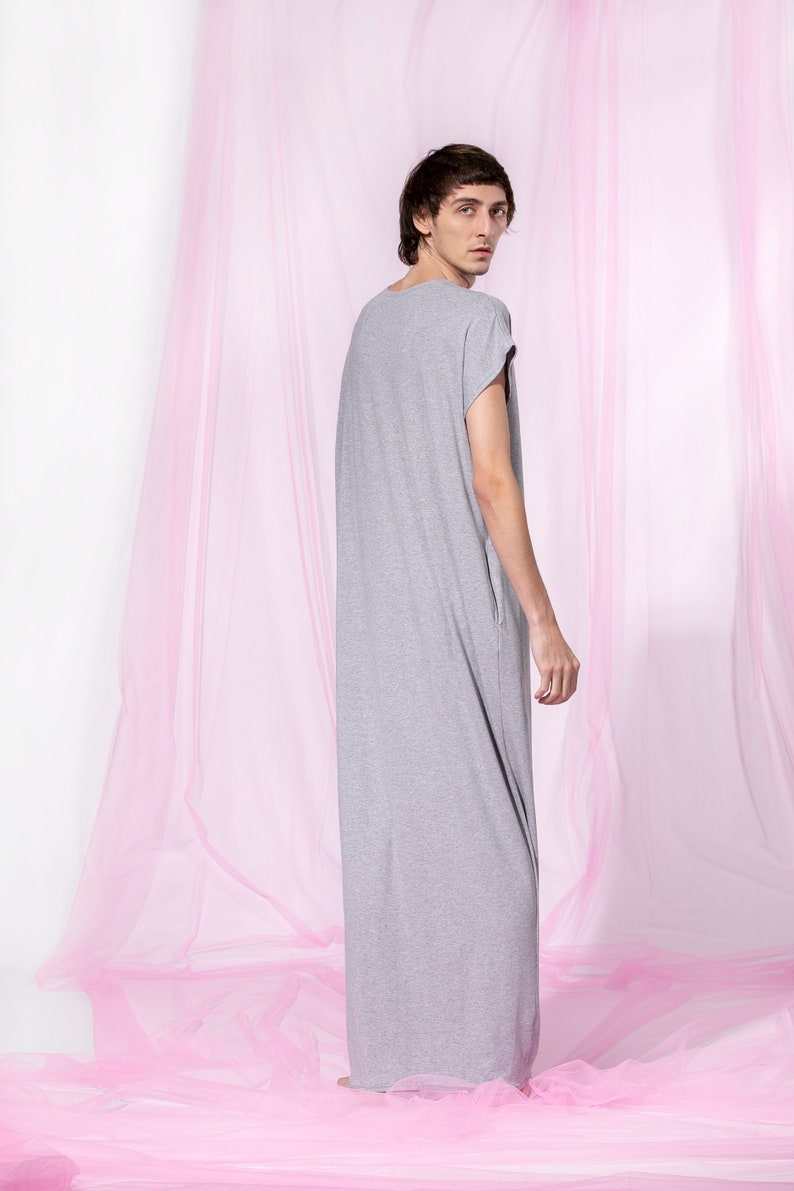 May include: A person wearing a long, gray, loose-fitting t-shirt dress standing in front of a pink sheer curtain.