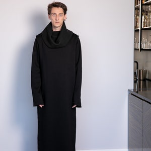May include: A black long-sleeved turtleneck sweater with a cowl neck. The sweater is long and falls to the ankles. The person wearing the sweater is standing in front of a white wall.