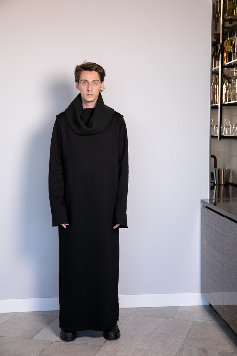 May include: A black long-sleeved turtleneck sweater with a cowl neck. The sweater is long and falls to the ankles. The person wearing the sweater is standing in front of a white wall.