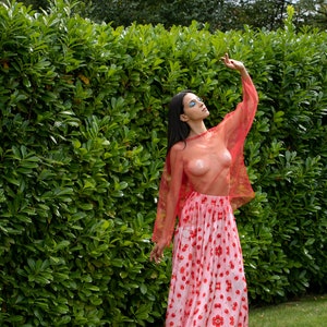 May include: A woman wearing a sheer red top with a cherry print skirt and pink sandals. She is standing in front of a green hedge.