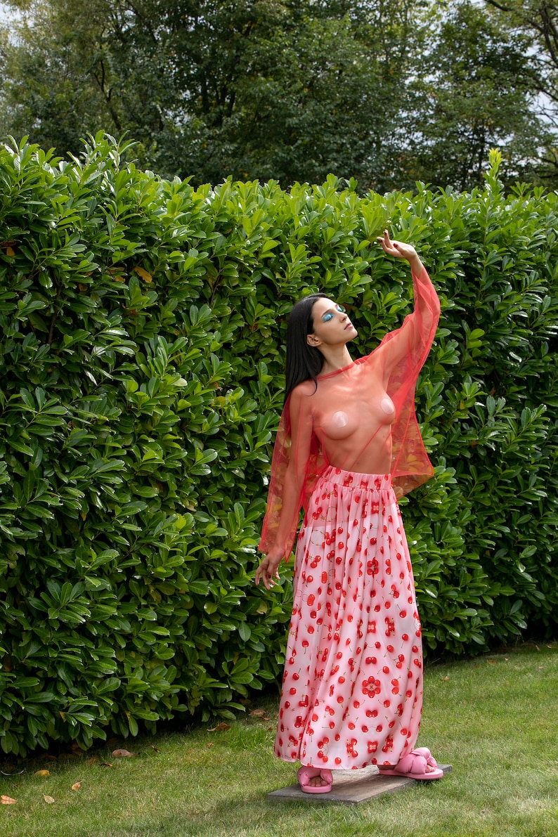 May include: A woman wearing a sheer red top with a cherry print skirt and pink sandals. She is standing in front of a green hedge.