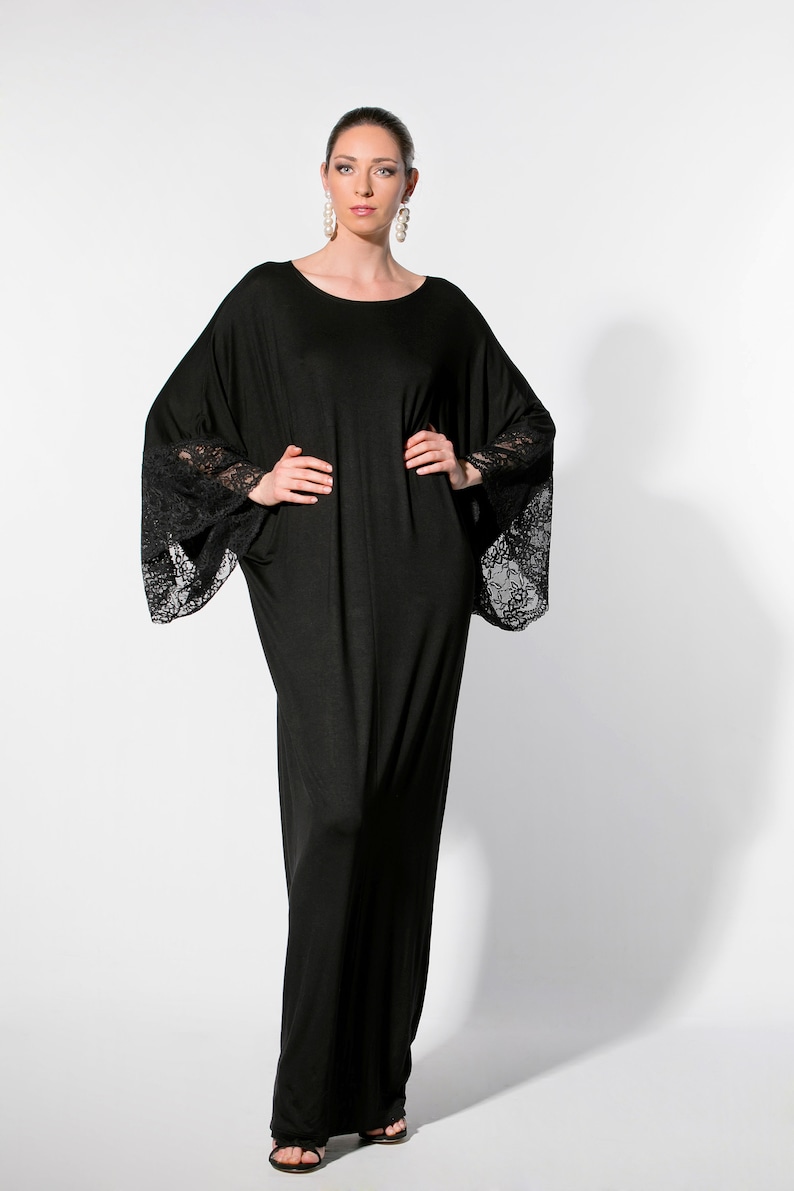 Puede incluir: Un vestido largo negro con mangas anchas y detalles de encaje en los pu&ntilde;os. El vestido tiene una silueta suelta y fluida y un cuello redondo. La modelo lleva pendientes colgantes grandes.