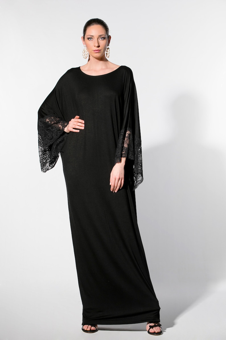 Puede incluir: Vestido negro largo con mangas anchas y pu&ntilde;os de encaje. El vestido tiene un escote redondo y se lleva con sandalias negras. La modelo lleva pendientes de perlas grandes.