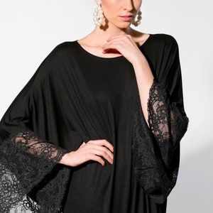Puede incluir: Vestido negro con mangas anchas y pu&ntilde;os de encaje negro. El vestido tiene un cuello redondo y una silueta suelta. La modelo lleva pendientes grandes con forma de perla.
