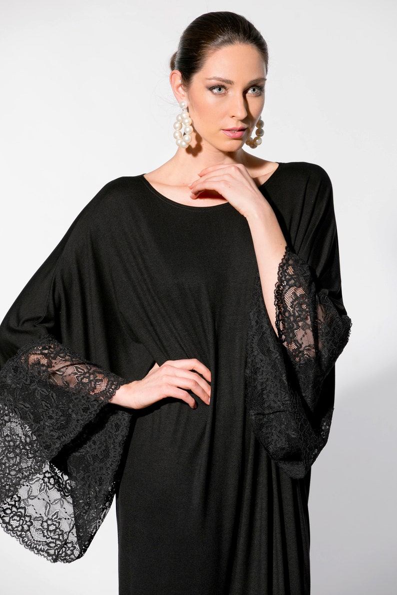 Puede incluir: Vestido negro con mangas anchas y pu&ntilde;os de encaje negro. El vestido tiene un cuello redondo y una silueta suelta. La modelo lleva pendientes grandes con forma de perla.