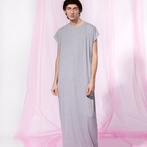 May include: A person wearing a long, gray, sleeveless shirt standing in front of a pink tulle backdrop.