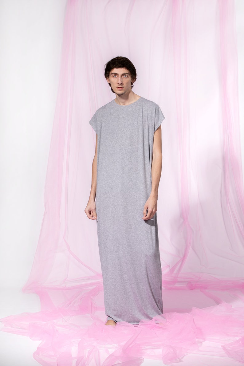 May include: A person wearing a long, gray, sleeveless shirt standing in front of a pink tulle backdrop.