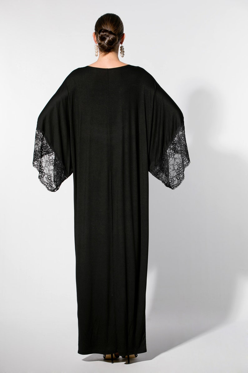 Puede incluir: Un vestido largo negro con mangas anchas y detalles de encaje en los pu&ntilde;os. El vestido tiene un dise&ntilde;o simple y elegante y una silueta fluida. La modelo est&aacute; de espaldas a la c&aacute;mara, mostrando la parte trasera del vestido.