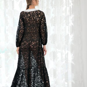 May include: A black lace maxi dress with a white collar and long sleeves. The dress has a flowing skirt and is shown on a model standing in front of a white curtain.
