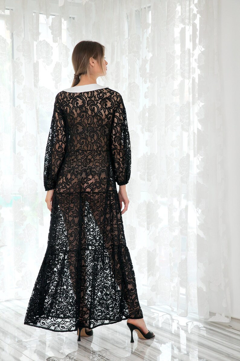 May include: A black lace maxi dress with a white collar and long sleeves. The dress has a flowing skirt and is shown on a model standing in front of a white curtain.