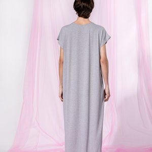 May include: A person wearing a long, gray, loose-fitting dress with short sleeves. The person is standing in front of a pink sheer curtain.