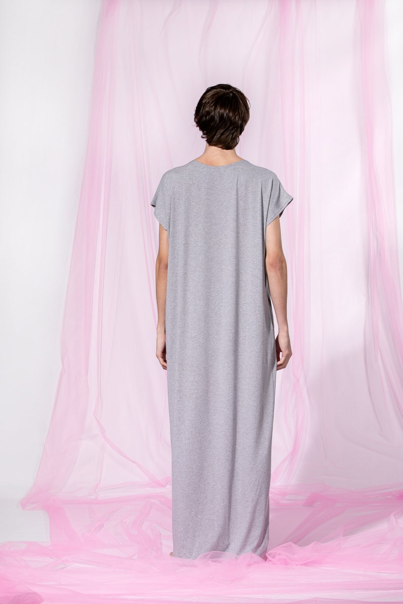 May include: A person wearing a long, gray, loose-fitting dress with short sleeves. The person is standing in front of a pink sheer curtain.